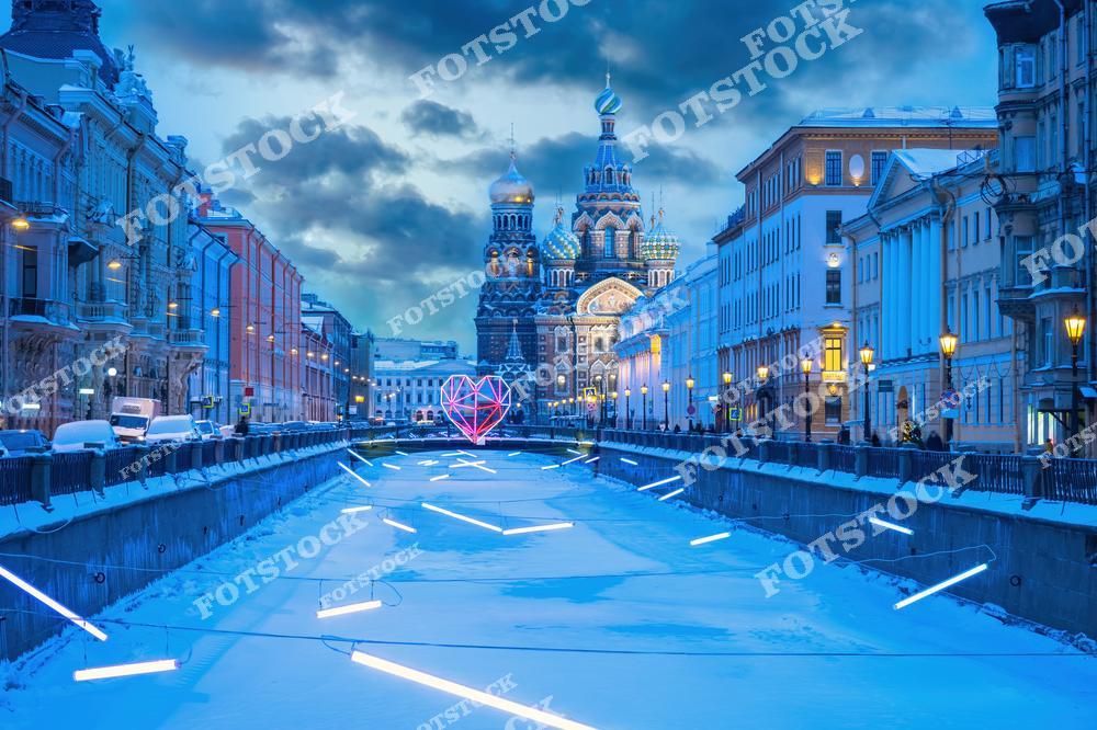 На фотографии изображена церковь Спаса на Крови в Санкт-Петербурге, окруженная снежной атмосферой и новогодними огнями. В центре композиции — яркая световая инсталляция в виде сердца, создающая контраст с историческим зданием. Вдоль канала и на улицах города видно много гирлянд и праздничных украшений, что придает Санкт-Петербургу особое очарование в зимний вечер.